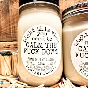 May include: Two glass jar candles with black lids and white labels that read "Light this when you need to CALM THE FUCK DOWN!". The candles are surrounded by matches with colorful tips. The candles are 16oz.