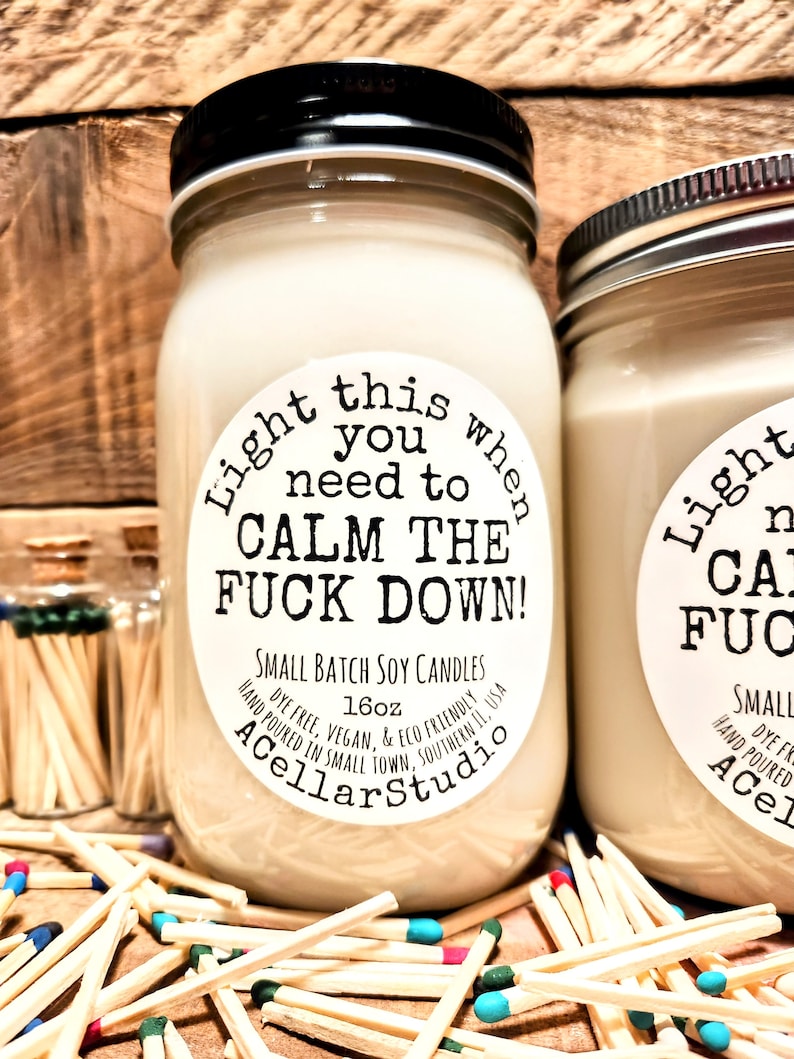 May include: Two glass jar candles with black lids and white labels that read "Light this when you need to CALM THE FUCK DOWN!". The candles are surrounded by matches with colorful tips. The candles are 16oz.