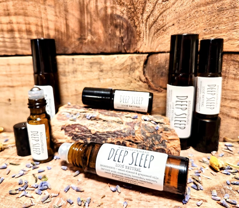 May include: A close-up of a brown glass roller bottle with a white label that reads "Deep Sleep 100% Natural". The label lists ingredients including sweet almond & grapeseed oils, lavender, chamomile, vetiver, and more. The bottle is surrounded by dried lavender flowers and other botanicals.