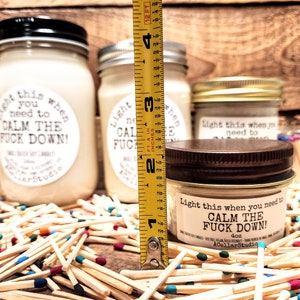 May include: Several soy candles in glass jars with black, silver, and bronze lids. The labels read "Light this when you need to CALM THE FUCK DOWN!". The candles are surrounded by matches with colorful tips. The tallest candle measures approximately 4 inches.