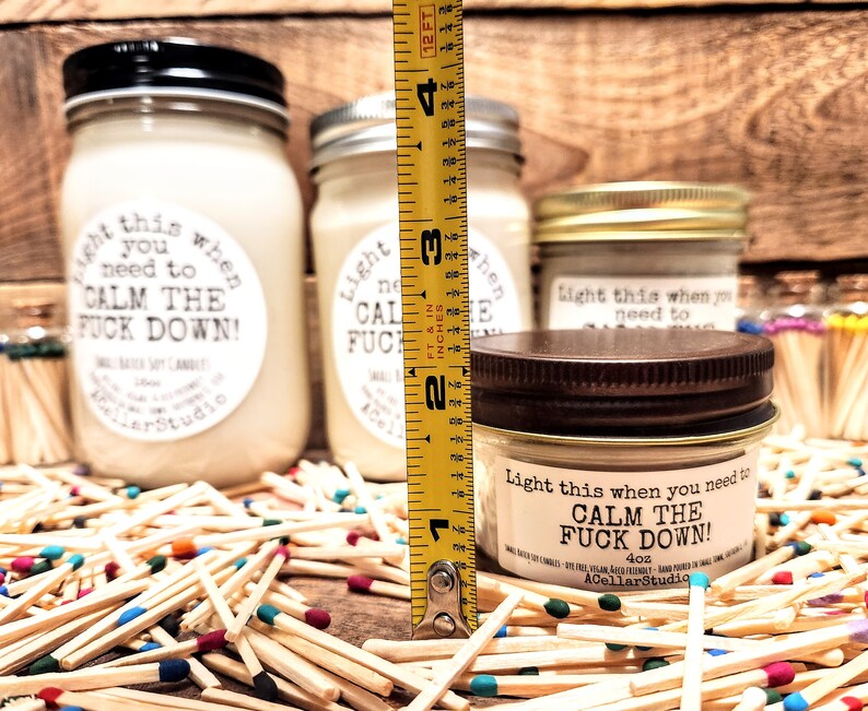 May include: Several soy candles in glass jars with black, silver, and bronze lids. The labels read "Light this when you need to CALM THE FUCK DOWN!". The candles are surrounded by matches with colorful tips. The tallest candle measures approximately 4 inches.
