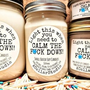 May include: Several small batch soy candles in glass jars with metal lids. The labels read "Light this when you need to CALM THE FUCK DOWN!" and "ACellarStudio." The candles are surrounded by colorful matchsticks.