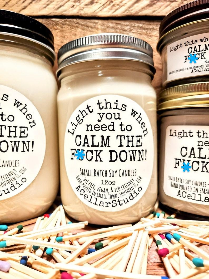 May include: Several small batch soy candles in glass jars with metal lids. The labels read "Light this when you need to CALM THE FUCK DOWN!" and "ACellarStudio." The candles are surrounded by colorful matchsticks.