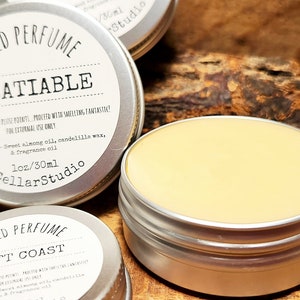 以下が含まれることがあります： 固形香水の銀色の缶が2つ。上の缶には「SOLID PERFUME INSATIABLE」とラベルが貼られており、使用方法と成分が記載されています。下の缶には「SOLID PERFUME LEFT COAST」とラベルが貼られています。