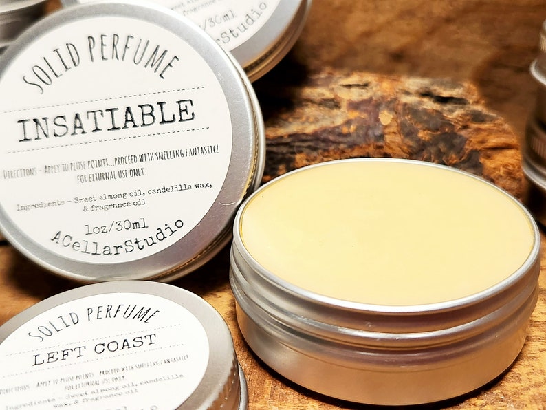 以下が含まれることがあります： 固形香水の銀色の缶が2つ。上の缶には「SOLID PERFUME INSATIABLE」とラベルが貼られており、使用方法と成分が記載されています。下の缶には「SOLID PERFUME LEFT COAST」とラベルが貼られています。