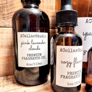 May include: Two glass bottles of fragrance oil with labels. The bottle on the left is labeled "ACellarStudio pink lavender clouds PREMIUM FRAGRANCE OIL 4oz/119ml". The bottle on the right is labeled "ACellarStudio cozy flann PREMIUM FRAGRANCE OIL 1oz/30ml".