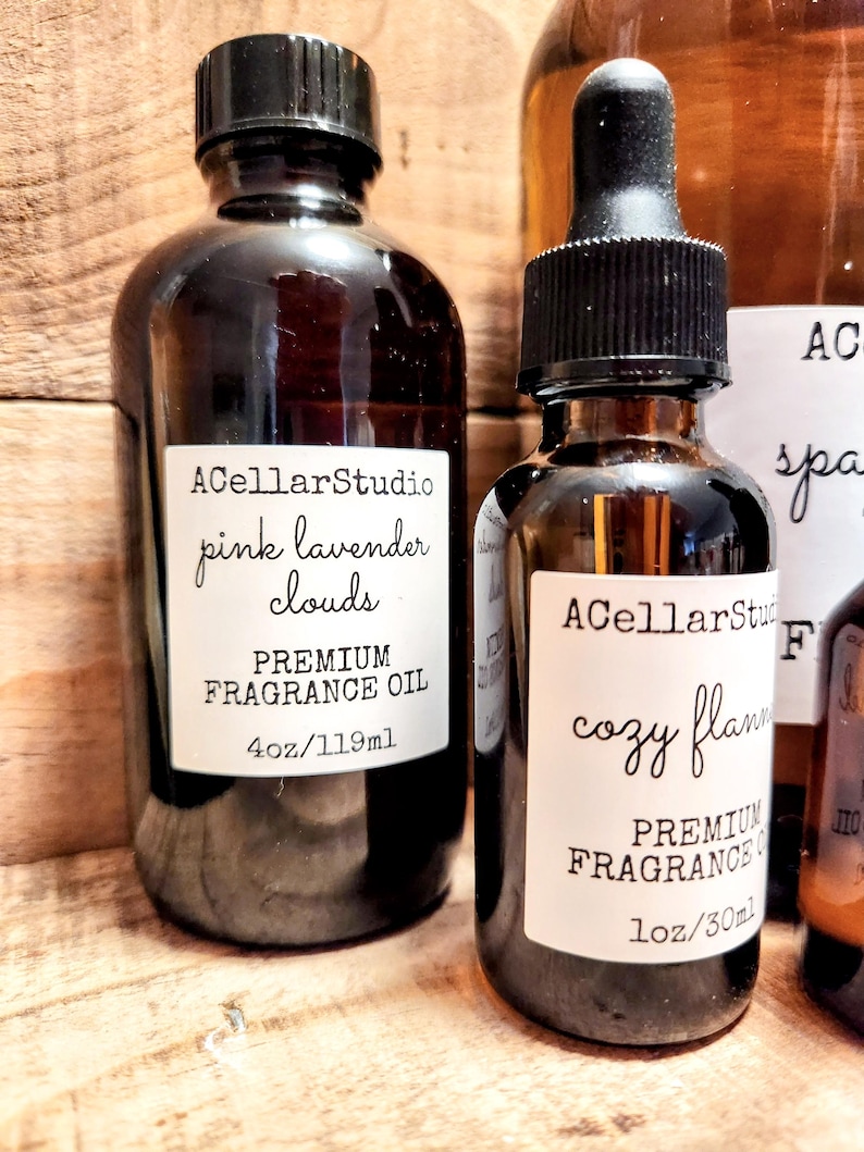 May include: Two glass bottles of fragrance oil with labels. The bottle on the left is labeled "ACellarStudio pink lavender clouds PREMIUM FRAGRANCE OIL 4oz/119ml". The bottle on the right is labeled "ACellarStudio cozy flann PREMIUM FRAGRANCE OIL 1oz/30ml".