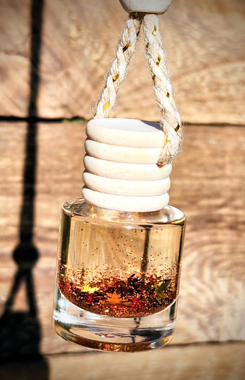 K&ouml;nnte beinhalten: Ein h&auml;ngender Auto-Lufterfrischer mit einer transparenten Glasflasche, gef&uuml;llt mit einer goldenen Fl&uuml;ssigkeit und Glitzer. Die Flasche ist mit Holzringen und einem geflochtenen Seil mit goldenen Akzenten versehen. Der Hintergrund ist eine Holzoberfl&auml;che.