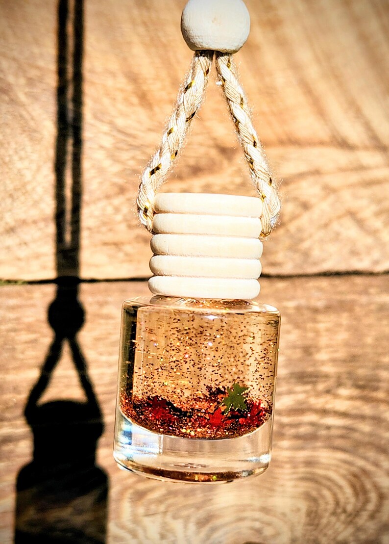 K&ouml;nnte beinhalten: Klares Glas-Lufterfrischer-Fl&auml;schchen f&uuml;r das Auto mit Holzdeckel und Aufh&auml;ngeschnur. Die Flasche enth&auml;lt eine Fl&uuml;ssigkeit mit Glitzer und kleinen roten und gr&uuml;nen Dekorationselementen. Der Hintergrund ist eine Holzoberfl&auml;che.