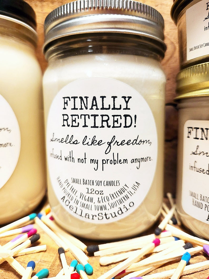 K&ouml;nnte beinhalten: Eine wei&szlig;e Sojawachskerze mit einem schwarz-wei&szlig;en Etikett, auf dem "FINALLY RETIRED! Smells like freedom, infused with not my problem anymore." steht. Das Etikett enth&auml;lt auch den Text "Small Batch Soy Candles, 12oz, Dye Free, Vegan, & Eco Friendly, Hand Poured in Small Town Southern IL, USA, A Cellar Studio".