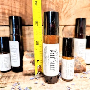 May include: Five glass roller bottles with black caps and white labels. The labels read "Deep Sleep" and "100% Natural". The bottles are arranged on a wooden surface with a yellow measuring tape in the background.