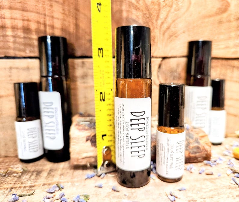 May include: Five glass roller bottles with black caps and white labels. The labels read "Deep Sleep" and "100% Natural". The bottles are arranged on a wooden surface with a yellow measuring tape in the background.