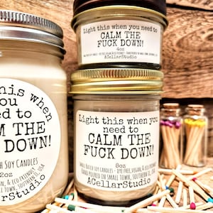 May include: Three glass jar candles with the text "Light this when you need to CALM THE FUCK DOWN!". The candles are surrounded by wooden matches with colorful tips. The candles are 4oz and 8oz.