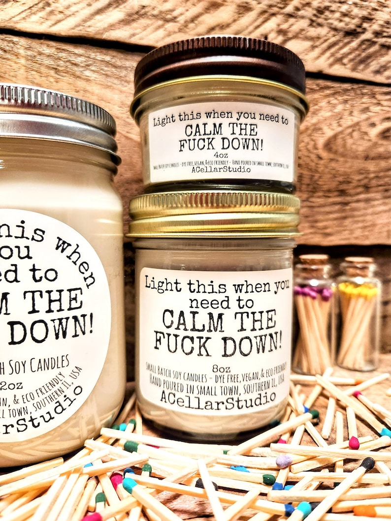 May include: Three glass jar candles with the text "Light this when you need to CALM THE FUCK DOWN!". The candles are surrounded by wooden matches with colorful tips. The candles are 4oz and 8oz.