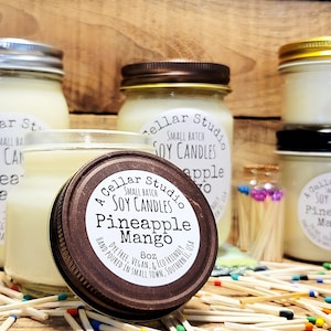 May include: A Cellar Studio small batch soy candle in a glass jar with a brown lid. The candle is labeled "Pineapple Mango" and is hand-poured, dye-free, vegan, and eco-friendly. The candle is surrounded by matchsticks.