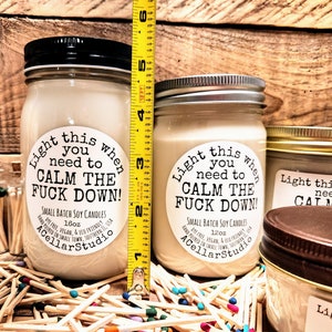 May include: Two soy candles in glass jars with black and silver lids. The labels read "Light this when you need to CALM THE FUCK DOWN!". The taller candle is approximately 5 inches tall. Matches are scattered around the candles.