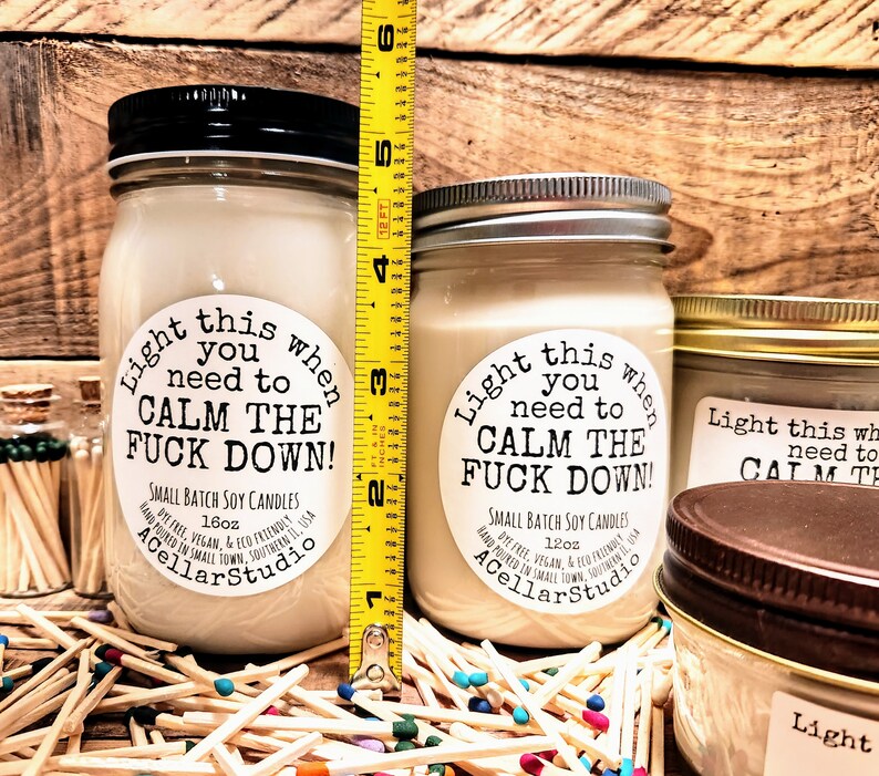 May include: Two soy candles in glass jars with black and silver lids. The labels read "Light this when you need to CALM THE FUCK DOWN!". The taller candle is approximately 5 inches tall. Matches are scattered around the candles.