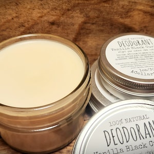 Puede incluir: Un tarro de vidrio lleno de un desodorante blanco cremoso, junto a una lata de plata con una etiqueta que dice "100% Natural Desodorante Vainilla Grosella Negra - Stink Naturally*" y "1 oz/30 ml ACellarStudio".
