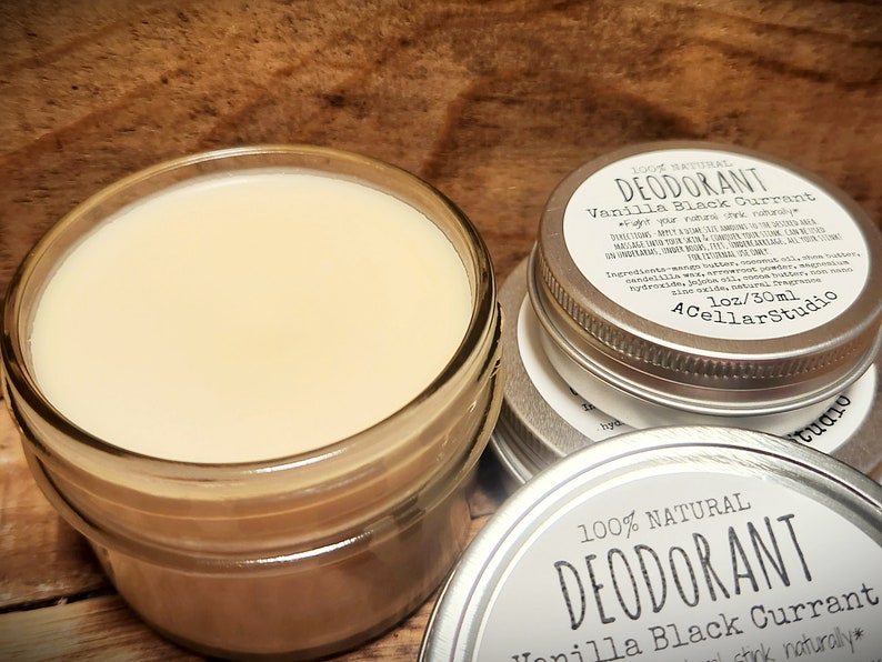 Puede incluir: Un tarro de vidrio lleno de un desodorante blanco cremoso, junto a una lata de plata con una etiqueta que dice "100% Natural Desodorante Vainilla Grosella Negra - Stink Naturally*" y "1 oz/30 ml ACellarStudio".