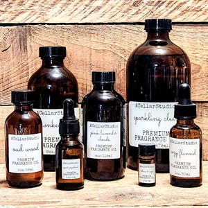 May include: A collection of amber glass bottles with droppers, containing premium fragrance oils. The bottles are labeled with names like "oud wood", "pink lavender clouds", "sparkling clean", and "cozy flannel". Each bottle has a label with the text "ACellarStudio" and "Premium Fragrance Oil".