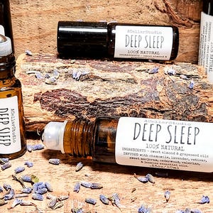 May include: A close-up of a brown glass roller bottle with a white label that reads "ACellarStudio DEEP SLEEP 100% NATURAL INGREDIENTS - sweet almond & grapeseed oils INFUSED with chamomile, lavender, vetiver, sweet marjoran, cedarwood, valerian, & jasmine essential oils. .33oz/10ml". The bottle is sitting on a wooden surface with lavender flowers scattered around it.