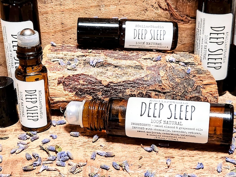 May include: A close-up of a brown glass roller bottle with a white label that reads "ACellarStudio DEEP SLEEP 100% NATURAL INGREDIENTS - sweet almond & grapeseed oils INFUSED with chamomile, lavender, vetiver, sweet marjoran, cedarwood, valerian, & jasmine essential oils. .33oz/10ml". The bottle is sitting on a wooden surface with lavender flowers scattered around it.