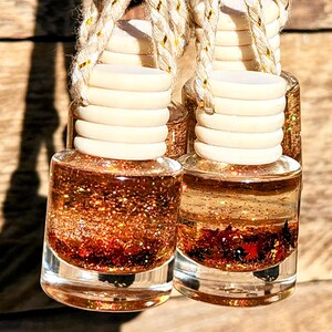 K&ouml;nnte beinhalten: Zwei kleine, klare Glasflaschen, gef&uuml;llt mit einer schimmernden, glitzernden Fl&uuml;ssigkeit in Kupfer- und Rott&ouml;nen. Jede Flasche hat einen hellen Holzdeckel und eine Schnur zum Aufh&auml;ngen. Der Hintergrund ist eine Holzoberfl&auml;che.