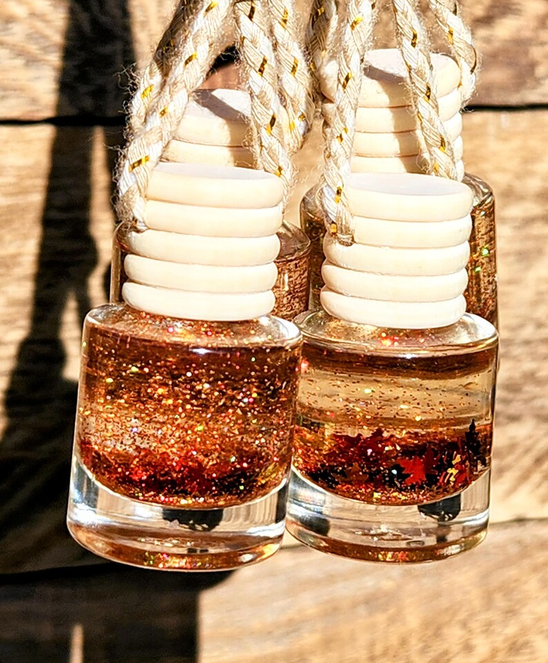 K&ouml;nnte beinhalten: Zwei kleine, klare Glasflaschen, gef&uuml;llt mit einer schimmernden, glitzernden Fl&uuml;ssigkeit in Kupfer- und Rott&ouml;nen. Jede Flasche hat einen hellen Holzdeckel und eine Schnur zum Aufh&auml;ngen. Der Hintergrund ist eine Holzoberfl&auml;che.