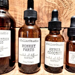K&ouml;nnte beinhalten: Mehrere braune Glasflaschen mit schwarzen Tropfern und wei&szlig;en Etiketten vor einem Holzgrund. Die Etiketten tragen die Aufschriften "Monkey Farts" und "Amber Coconut" mit dem Text "scented oil diffuser REFILL".