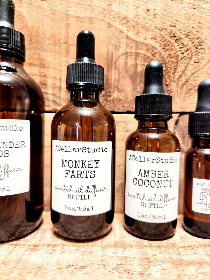 K&ouml;nnte beinhalten: Mehrere braune Glasflaschen mit schwarzen Tropfern und wei&szlig;en Etiketten vor einem Holzgrund. Die Etiketten tragen die Aufschriften "Monkey Farts" und "Amber Coconut" mit dem Text "scented oil diffuser REFILL".