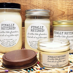K&ouml;nnte beinhalten: Drei Glasbeh&auml;lter mit wei&szlig;en Kerzen. Jedes Gef&auml;&szlig; hat ein wei&szlig;es Etikett mit dem Text "FINALLY RETIRED! Smells like freedom, infused with not my problem anymore." Die Beh&auml;lter stehen auf einer Oberfl&auml;che, die mit bunten Streichh&ouml;lzern bedeckt ist.