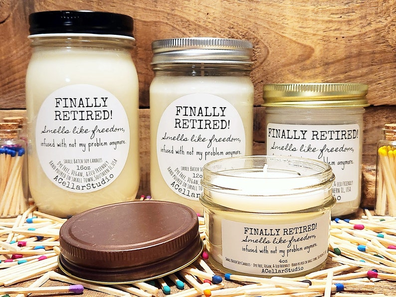K&ouml;nnte beinhalten: Drei Glasbeh&auml;lter mit wei&szlig;en Kerzen. Jedes Gef&auml;&szlig; hat ein wei&szlig;es Etikett mit dem Text "FINALLY RETIRED! Smells like freedom, infused with not my problem anymore." Die Beh&auml;lter stehen auf einer Oberfl&auml;che, die mit bunten Streichh&ouml;lzern bedeckt ist.