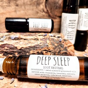 May include: A brown glass roller bottle with a white label that reads "DEEP SLEEP 100% NATURAL" and lists the ingredients. The bottle is sitting on a wooden surface with purple flowers.