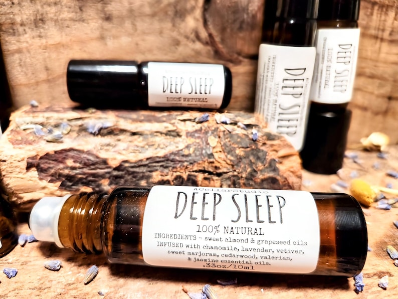 May include: A brown glass roller bottle with a white label that reads "DEEP SLEEP 100% NATURAL" and lists the ingredients. The bottle is sitting on a wooden surface with purple flowers.