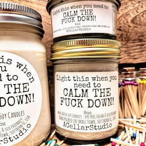 May include: Three glass jars of soy candles with metal lids, labeled "Light this when you need to CALM THE FUCK DOWN!". The jars are surrounded by wooden matches. The labels also include the size of the candle, 4oz and 8oz, and the name of the company, ACellarStudio.