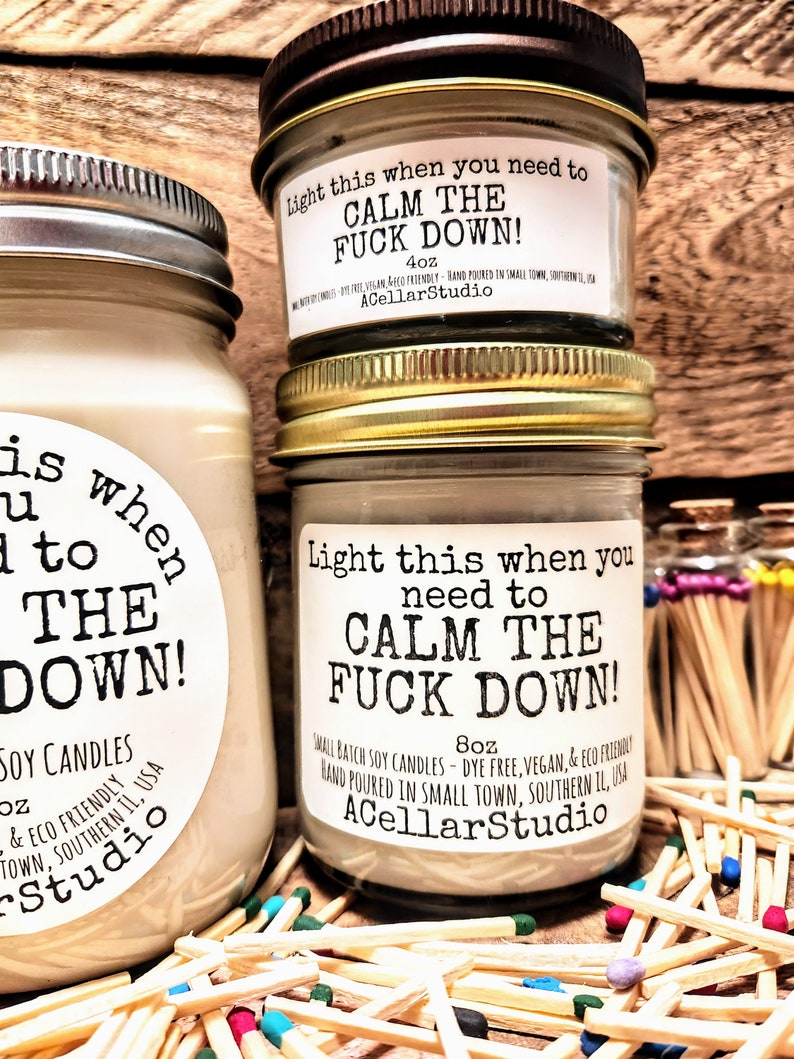 May include: Three glass jars of soy candles with metal lids, labeled "Light this when you need to CALM THE FUCK DOWN!". The jars are surrounded by wooden matches. The labels also include the size of the candle, 4oz and 8oz, and the name of the company, ACellarStudio.