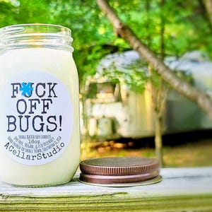 May include: A glass jar candle with a white label that says "F*CK OFF BUGS!" in black lettering. The candle is 16oz and is hand-poured in small town, Southern USA. The label also says "A Cellar Studio".