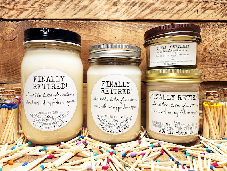 K&ouml;nnte beinhalten: Drei Glasbeh&auml;lter mit Sojawachskerzen. Jedes Glas hat ein wei&szlig;es Etikett mit dem Text "FINALLY RETIRED! Smells like freedom, infused with not my problem anymore." Die Gl&auml;ser stehen auf einer Holzoberfl&auml;che, umgeben von bunten Streichh&ouml;lzern.
