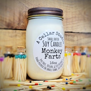 May include: A large soy candle in a mason jar with a brown lid, labeled "Monkey Farts." The label also reads "A Cellar Studio, Small Batch Soy Candles." Several small glass bottles of matches with colored tips are in the background. The candle is 16 oz.