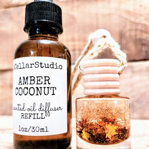 K&ouml;nnte beinhalten: Eine braune Glasflasche mit schwarzer Pipette und ein Auto-Lufterfrischer. Die Flasche tr&auml;gt das Etikett "Amber Coconut" und "Duft&ouml;l Diffusor Nachf&uuml;llung, 30ml". Der Lufterfrischer ist mit Glitzer und dekorativen Bl&auml;ttern gef&uuml;llt.