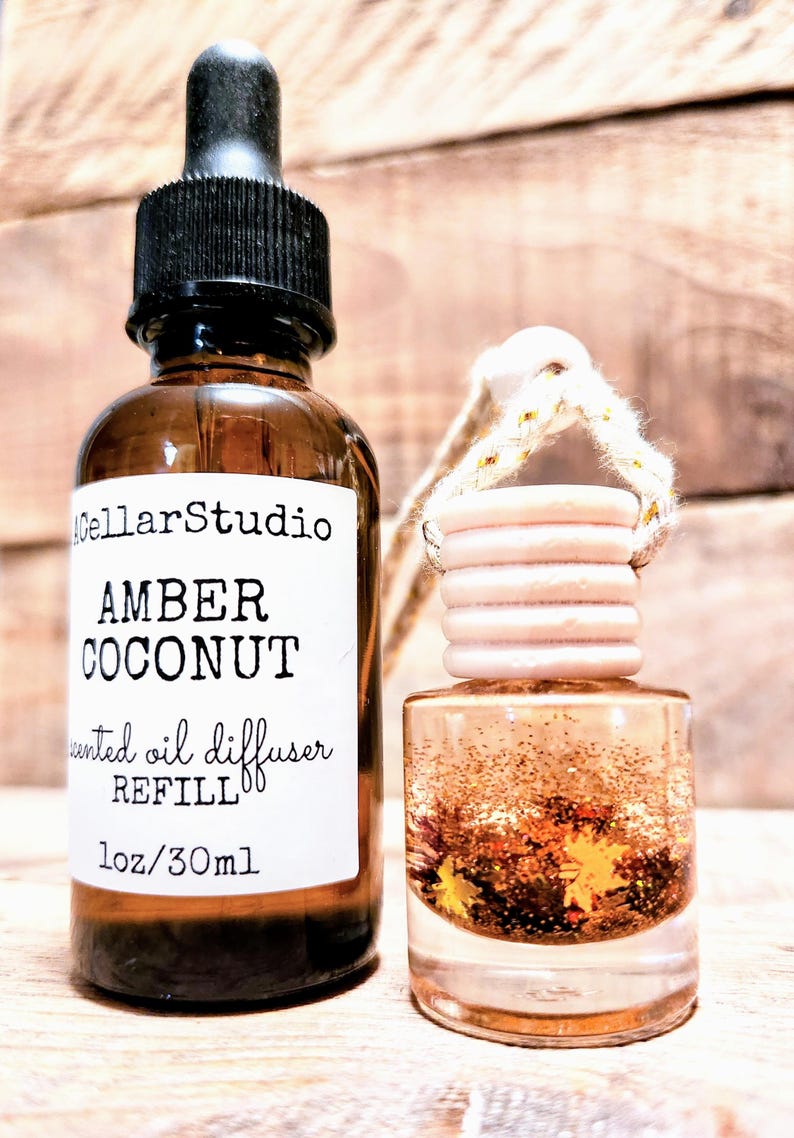 K&ouml;nnte beinhalten: Eine braune Glasflasche mit schwarzer Pipette und ein Auto-Lufterfrischer. Die Flasche tr&auml;gt das Etikett "Amber Coconut" und "Duft&ouml;l Diffusor Nachf&uuml;llung, 30ml". Der Lufterfrischer ist mit Glitzer und dekorativen Bl&auml;ttern gef&uuml;llt.