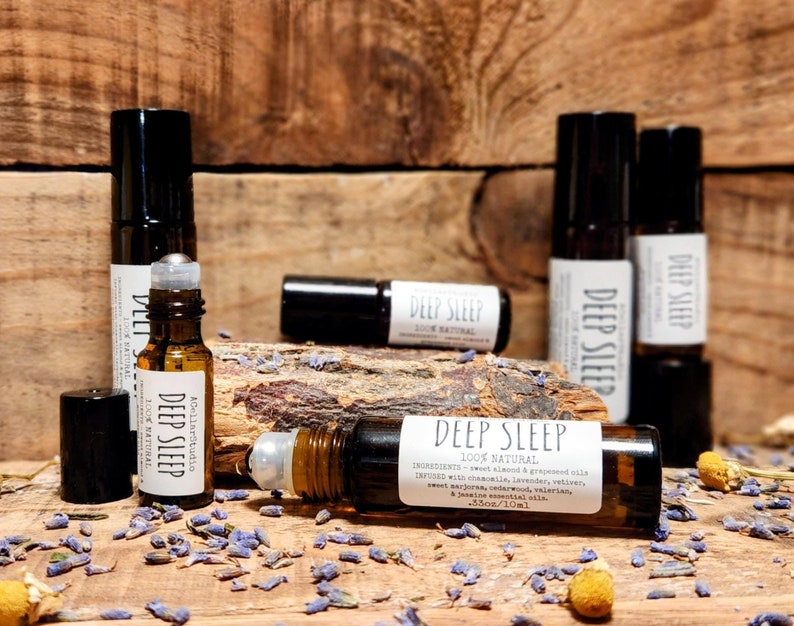 May include: A close-up of a brown glass roller bottle with a black cap and a white label that reads "Deep Sleep 100% Natural". The bottle is surrounded by dried lavender flowers and other botanicals. The label also lists the ingredients, which include chamomile, lavender, vetiver, sweet marjoram, cedarwood, and valerian. The bottle is 3.5 oz/10 ml.