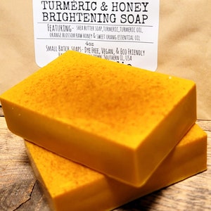 May include: Two bars of bright yellow turmeric and honey soap, stacked on top of each other. The soap is labeled "100% Natural Turmeric & Honey Brightening Soap" and "4oz Small Batch Soaps - Dye Free, Vegan, & Eco Friendly"