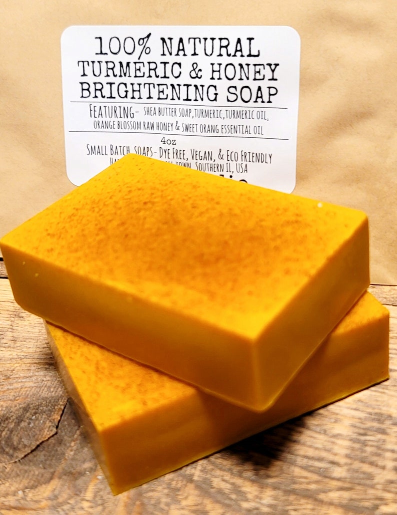 May include: Two bars of bright yellow turmeric and honey soap, stacked on top of each other. The soap is labeled "100% Natural Turmeric & Honey Brightening Soap" and "4oz Small Batch Soaps - Dye Free, Vegan, & Eco Friendly"