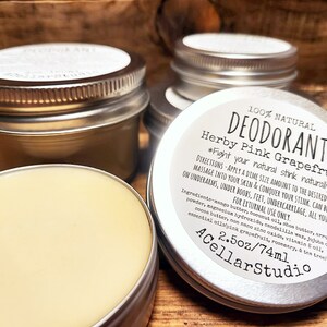 May include: A silver tin with a white label that reads "100% NATURAL DEODORANT Herby Pink Grapefruit *Fight your natural stink naturally*" with directions for use and ingredients listed. The tin contains a light yellow deodorant cream and is labeled "2.5oz/74ml ACellarStudio".