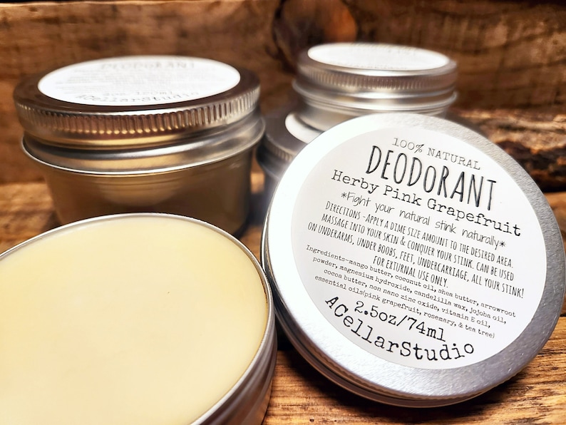 May include: A silver tin with a white label that reads "100% NATURAL DEODORANT Herby Pink Grapefruit *Fight your natural stink naturally*" with directions for use and ingredients listed. The tin contains a light yellow deodorant cream and is labeled "2.5oz/74ml ACellarStudio".