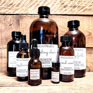 May include: A collection of amber glass bottles with black lids containing premium fragrance oils. The bottles are labeled with the text "A Cellar Studio" and various scents, including "Sparkling Clean", "Cozy Flannel", "Pink Grapefruit", "Oud Wood", and "Baby Farts". The bottles are arranged on a wooden surface.