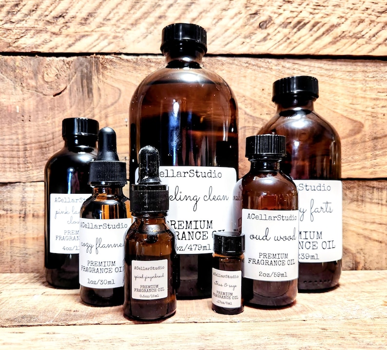 May include: A collection of amber glass bottles with black lids containing premium fragrance oils. The bottles are labeled with the text "A Cellar Studio" and various scents, including "Sparkling Clean", "Cozy Flannel", "Pink Grapefruit", "Oud Wood", and "Baby Farts". The bottles are arranged on a wooden surface.