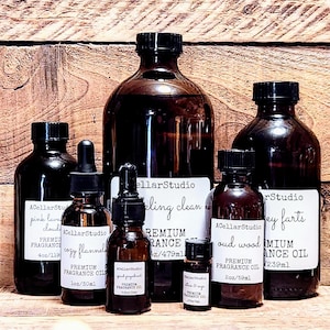 May include: A collection of seven amber glass bottles with dropper tops, containing various essential oils. The bottles are labeled with the brand name "ACellarStudio" and the names of the scents, including "pink lavender cloud", "cozy flannel", "bling clean", "oud wood", and "grey farts". The bottles are arranged on a wooden surface.