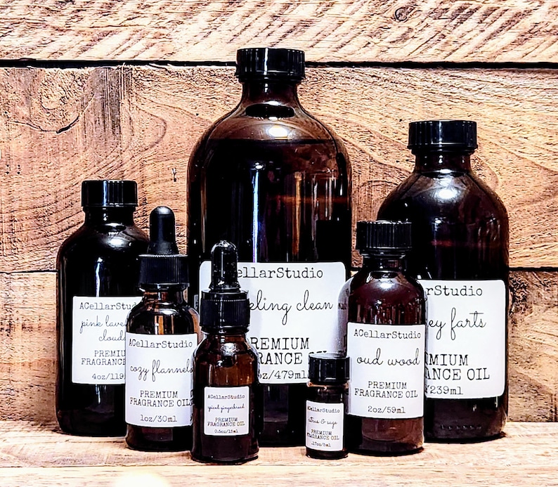 May include: A collection of seven amber glass bottles with dropper tops, containing various essential oils. The bottles are labeled with the brand name "ACellarStudio" and the names of the scents, including "pink lavender cloud", "cozy flannel", "bling clean", "oud wood", and "grey farts". The bottles are arranged on a wooden surface.
